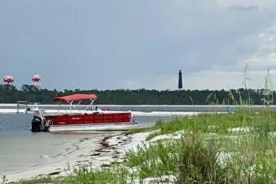 A red pontoon boat is anchored along a quiet sandbar in the Santa Rosa Sound near Pensacola Beach, with sea oats in the foreground and the Pensacola Lighthouse rising in the distance under a calm, overcast sky.