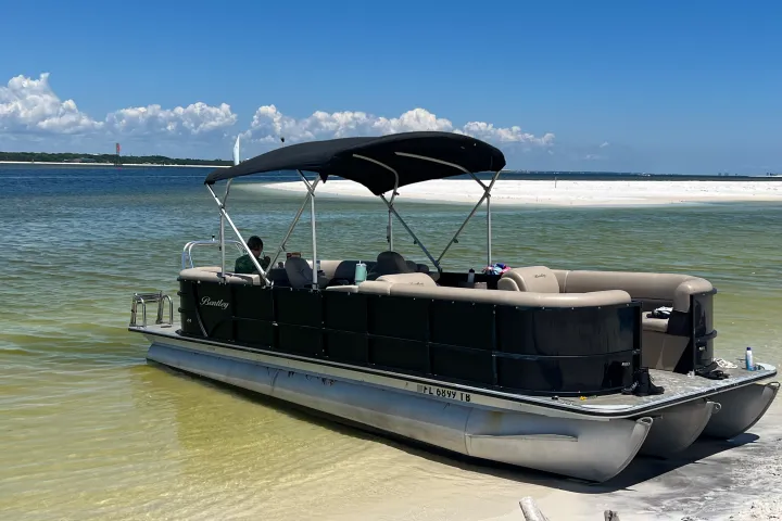 A black-canopy pontoon boat rests in shallow emerald water near a white sandbar in the Santa Rosa Sound, with calm Gulf Coast waters and bright blue skies surrounding the boat near Pensacola Beach.