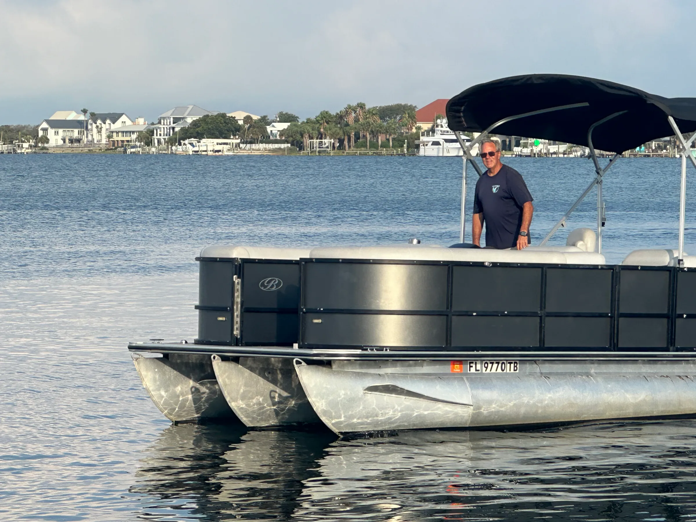 A pontoon boat cruising on calm water in Pensacola with a captain standing at the helm and waterfront homes in the background.