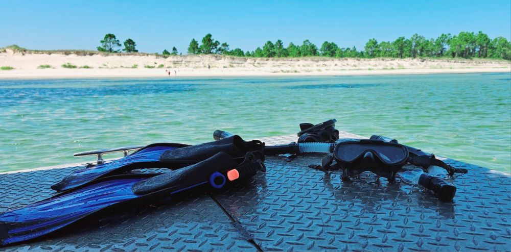 Snorkel fins, mask, and snorkel rest on the deck of a boat, overlooking calm emerald waters and white sand dunes along the Santa Rosa Sound near Pensacola Beach.