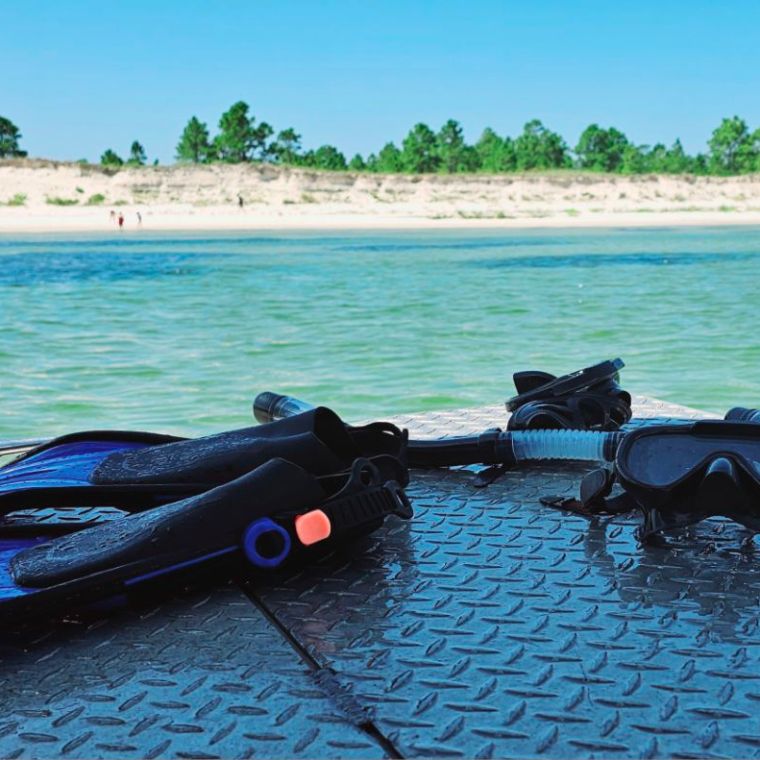 Snorkel fins, mask, and snorkel rest on the deck of a boat, overlooking calm emerald waters and white sand dunes along the Santa Rosa Sound near Pensacola Beach.