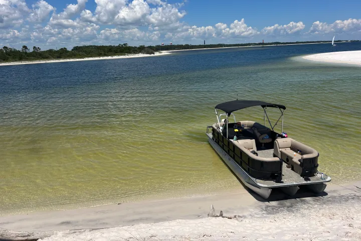 A pontoon boat pulled up to a shallow sandbar with clear green water and white sand in the Santa Rosa Sound near Pensacola Beach, Florida.