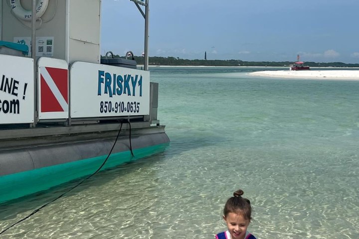 A child playing in shallow, clear water near Sand Island in the Santa Rosa Sound beside a Frisky Mermaid tour boat in Pensacola Beach, Florida.