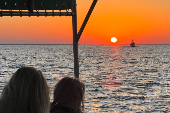 Passengers watch the sunset from a boat on Pensacola Bay with glowing orange skies reflecting across Santa Rosa Sound and the calm evening water.