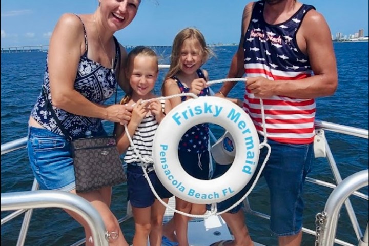 Family smiles on the bow of a tour boat in Pensacola Bay, holding a Frisky Mermaid life ring during a dolphin cruise.
