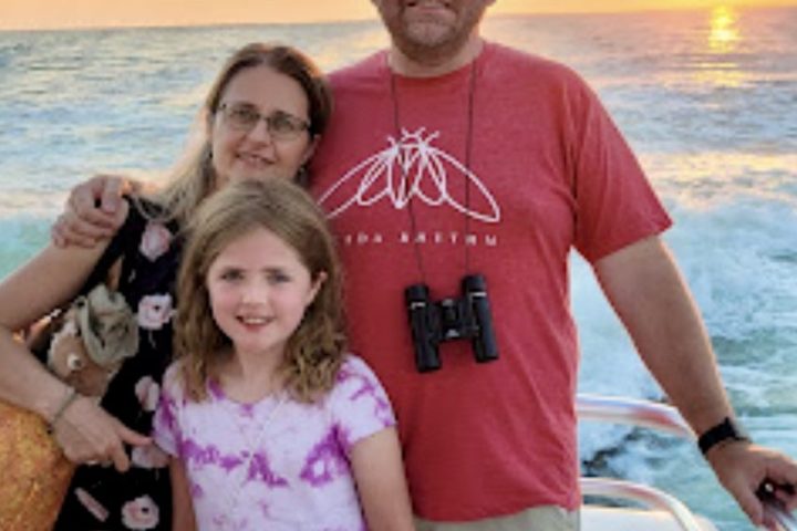Family smiles aboard a Frisky Mermaid sunset cruise on the Santa Rosa Sound with golden skies behind them.