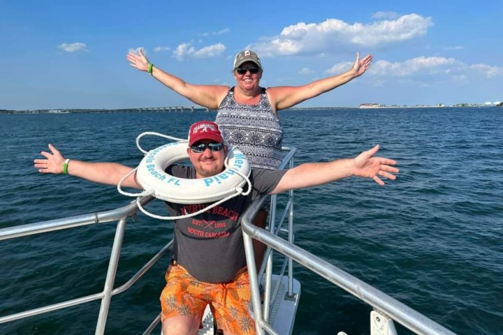 Smiling couple spreads their arms and cheer at the bow during a Frisky Mermaid dolphin cruise on the Santa Rosa Sound