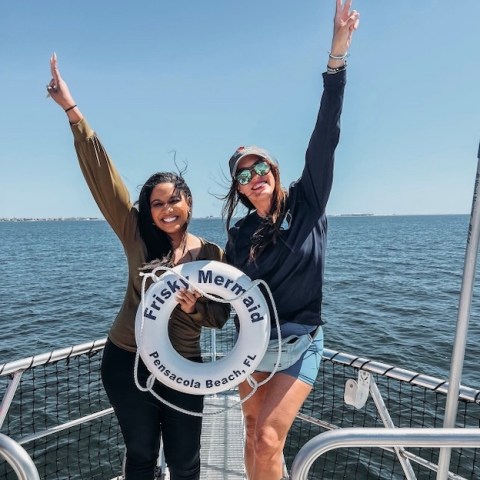 Captain Ally and Shan Bailey with NOLA news posing with a Frisky Mermaid life ring on a dolphin cruise boat in Santa Rosa Sound near Pensacola Beach, Florida. now holding a life ring labeled 'Frisky Mermaid', with water in the background.