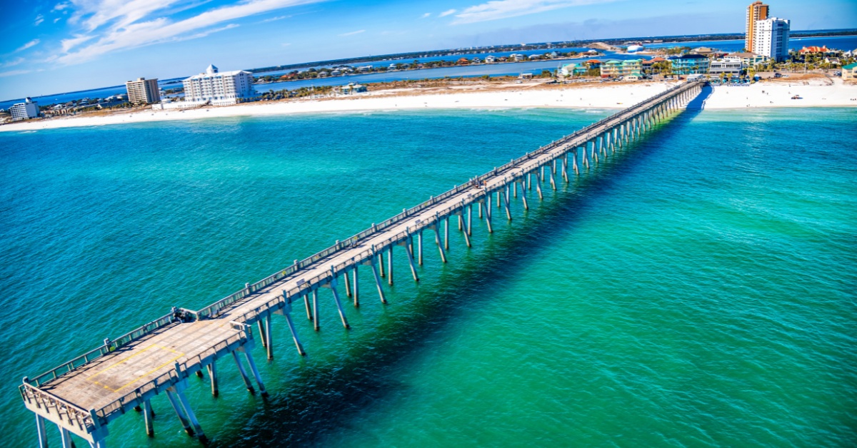 A long pier extends over turquoise ocean toward a distant sandy beach with buildings and blue sky.