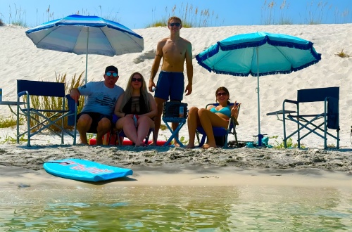 Four people on beach chairs under blue umbrellas, sitting on sand dunes.