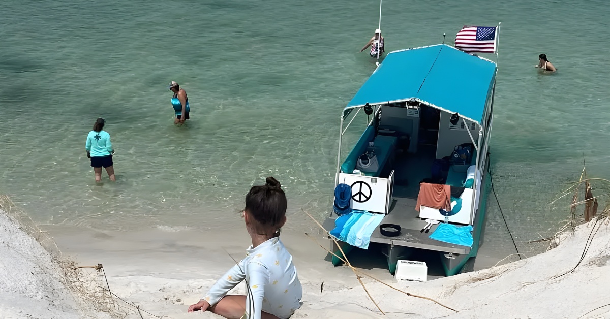 People relax near a small boat on a sandy beach with clear water.